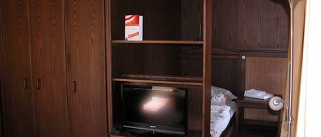A compact room with a wooden wardrobe, a television on a wooden stand, a small bedside table with a lamp, and a bed with a floral bedspread.