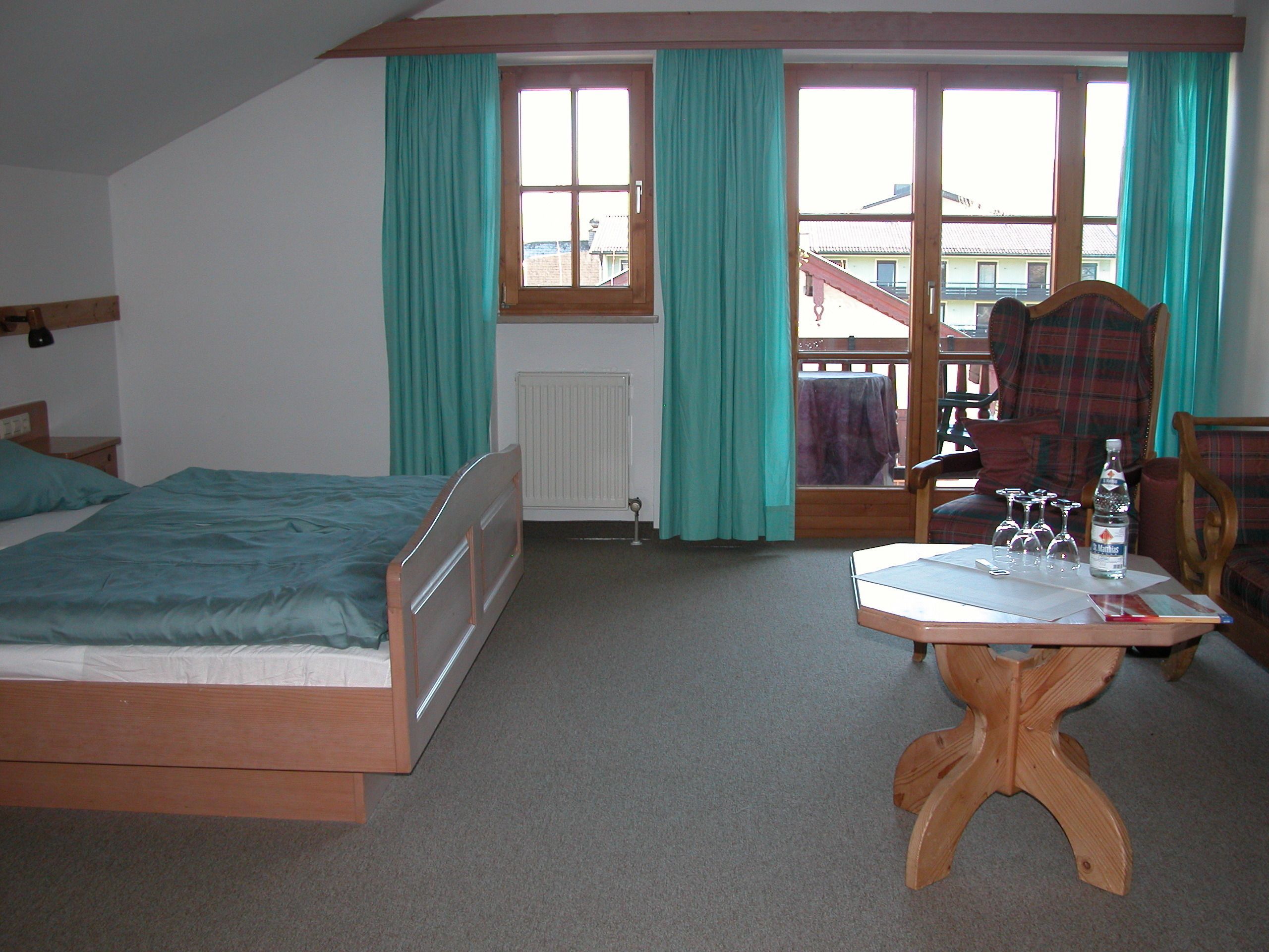 A bedroom with a bed, a wooden table with glasses and a bottle, and a view of the outside through a window.