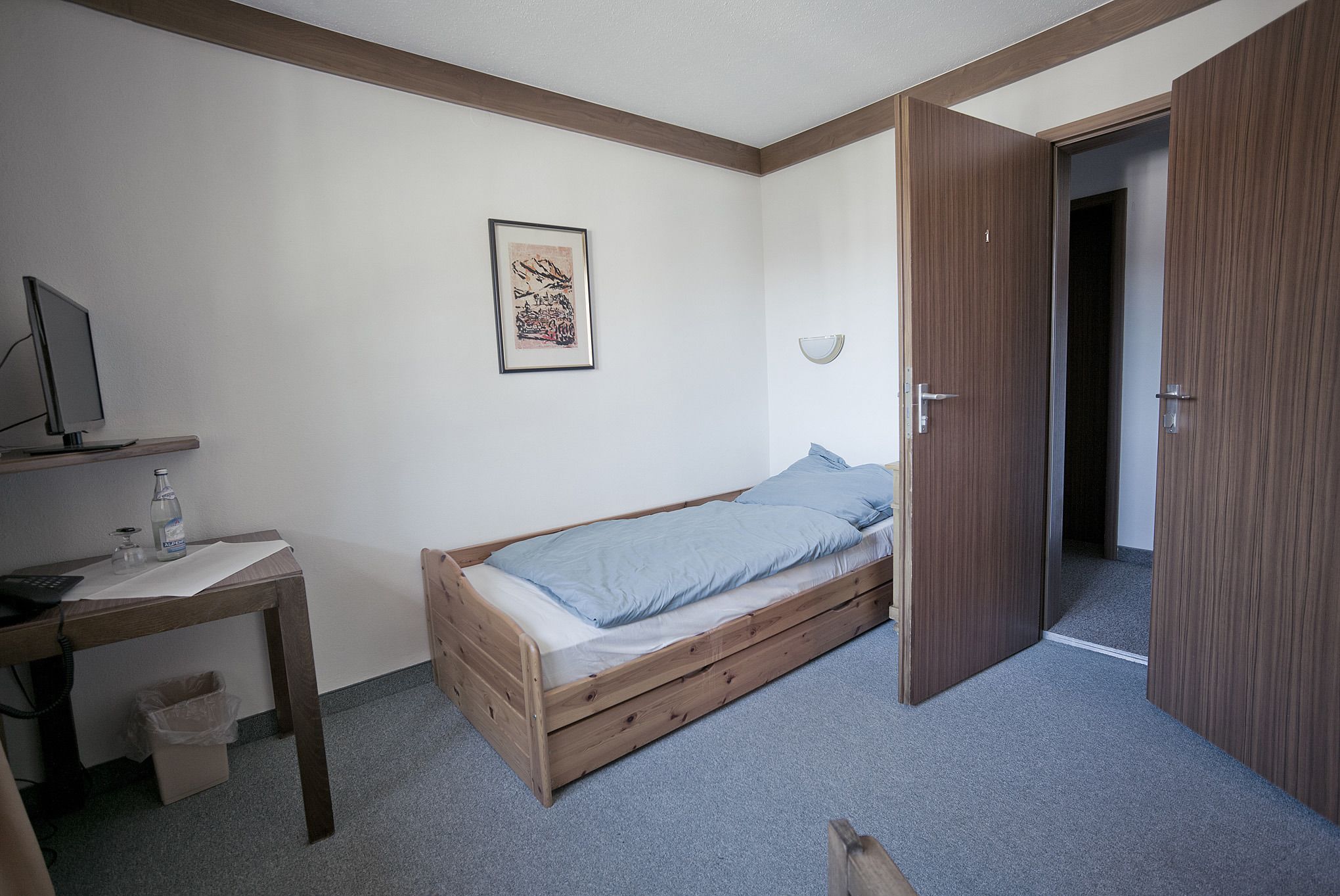 Ein Schlafzimmer mit einem Einzelbett, einem Holztisch mit einem Computer, einer kleinen Mülltonne und einer Tür zu einem anderen Raum.