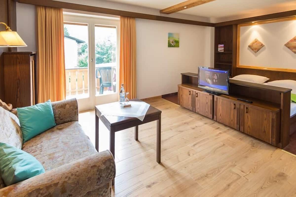 A cozy living room with a sofa, a table, a TV on a wooden cabinet, and a balcony with curtains.