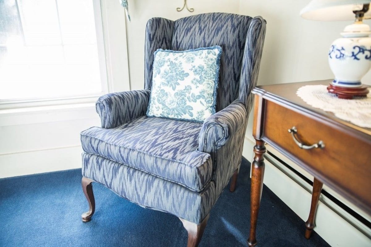 A chair with a patterned fabric and a floral pillow, positioned next to a wooden side table with a lamp.