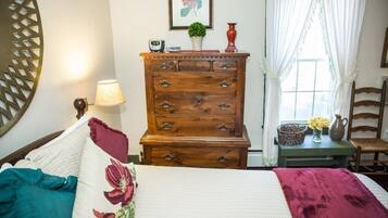A bedroom with a wooden dresser, a bed with floral bedding, a lamp, a window with curtains, and a chair.