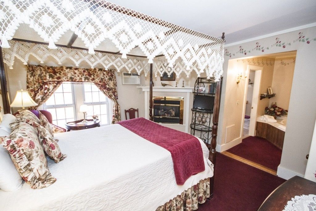 A bedroom with a large bed, floral curtains, a lace canopy, a fireplace, and a small bathroom.