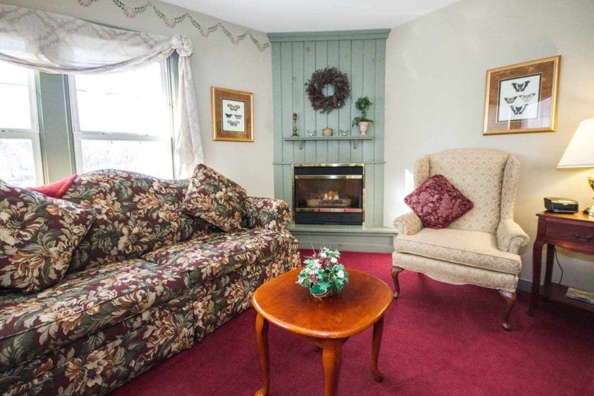 A living room with a floral sofa, a cream armchair, a small wooden table with a vase, a fireplace, and framed artwork on the walls.