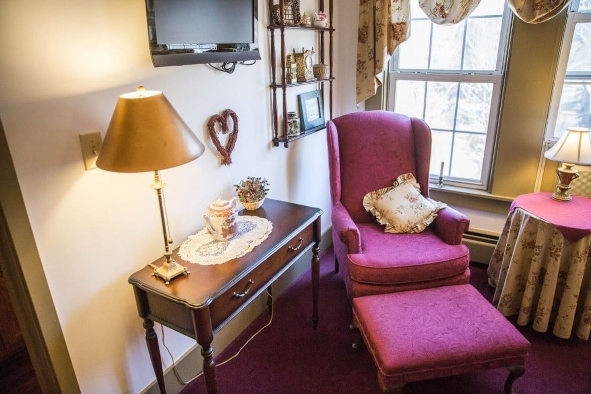 A cozy room with a wooden desk, a pink armchair, a small table with a lamp, and a television on the wall.
