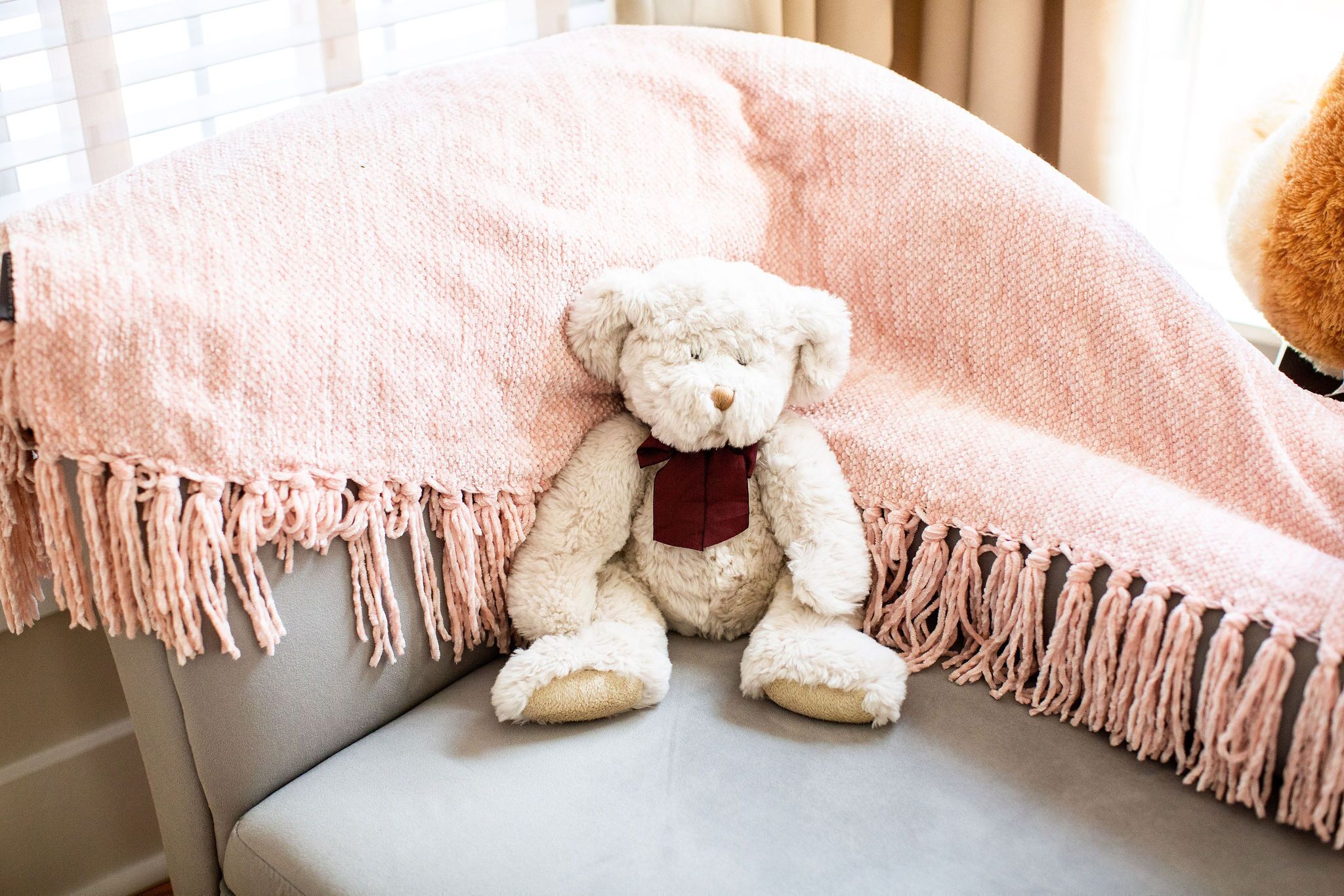 A white teddy bear with a red bow sitting on a grey couch covered with a pink fringed blanket.