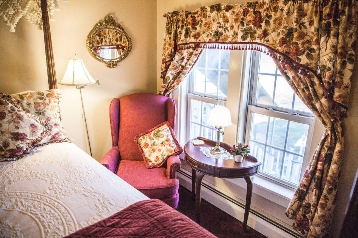 A bedroom with a bed, a red armchair, a small table with a lamp, and a window with floral curtains.