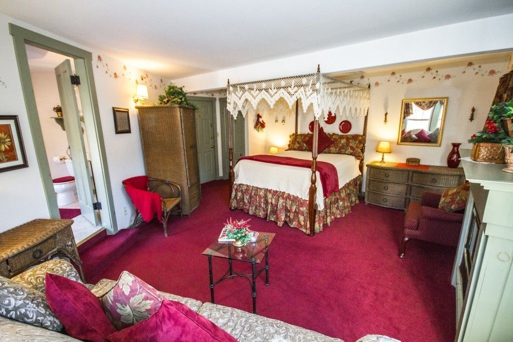 A traditional bedroom with a four-poster bed, a red carpet, a wooden wardrobe, a small table with a vase, and a red armchair.