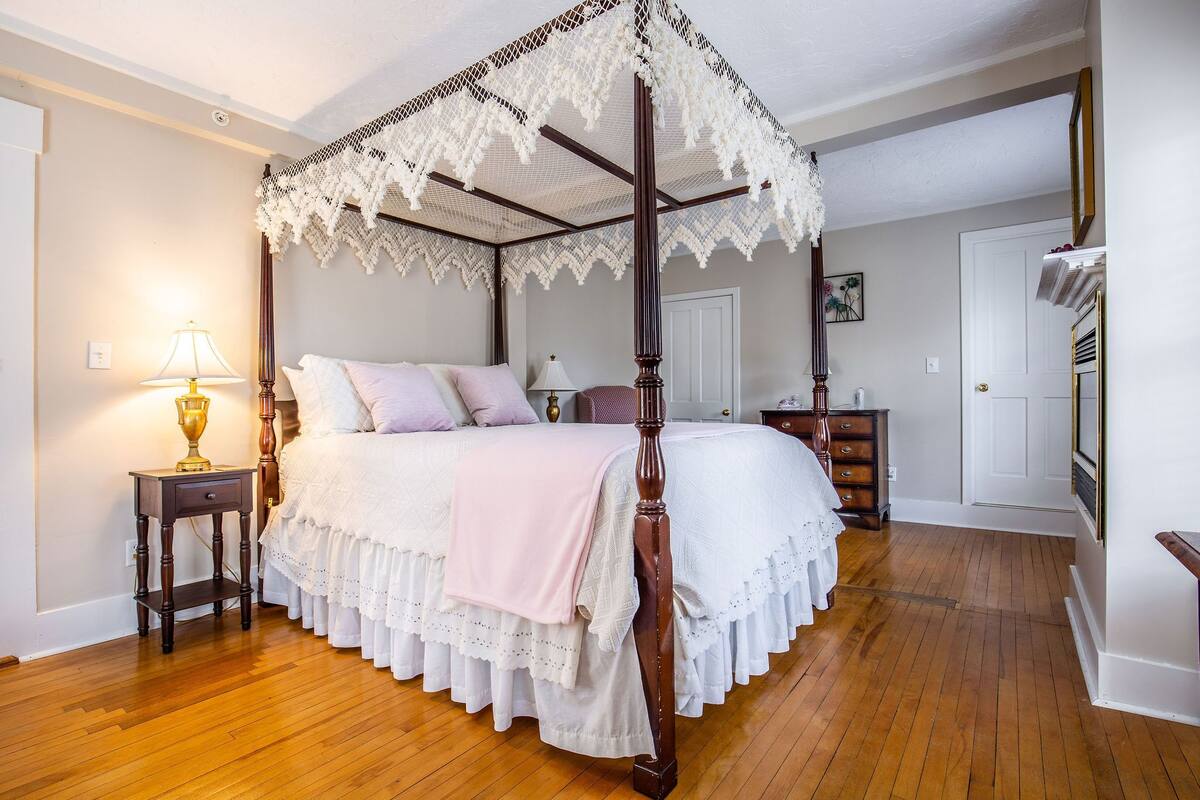 A four-poster canopy bed with a lace canopy, a bedside table with a lamp, and a wooden floor.