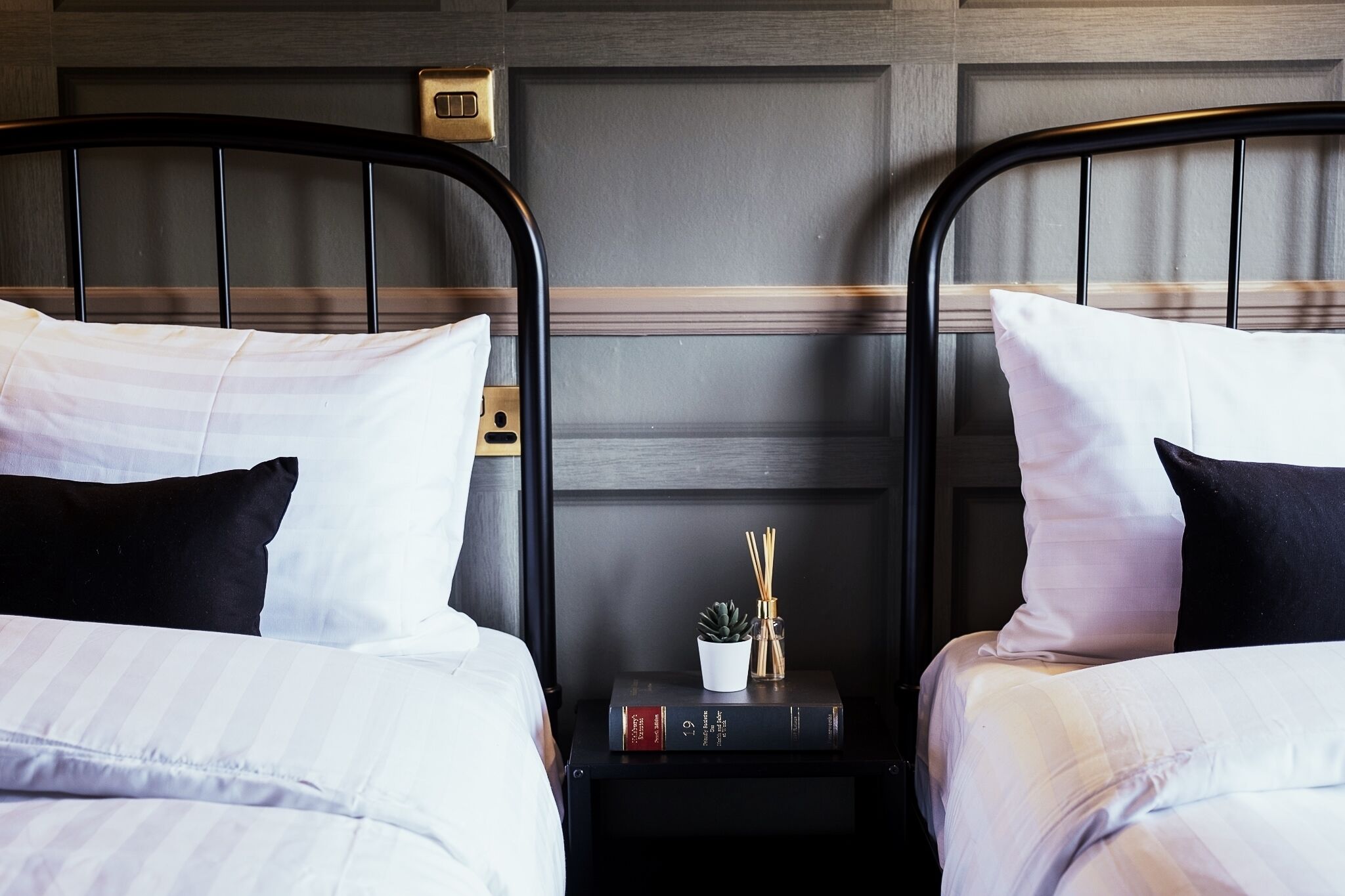 A hotel room with two beds, each with white bedding and black headboards. There is a small table between the beds with a book, a plant, and a decorative item.
