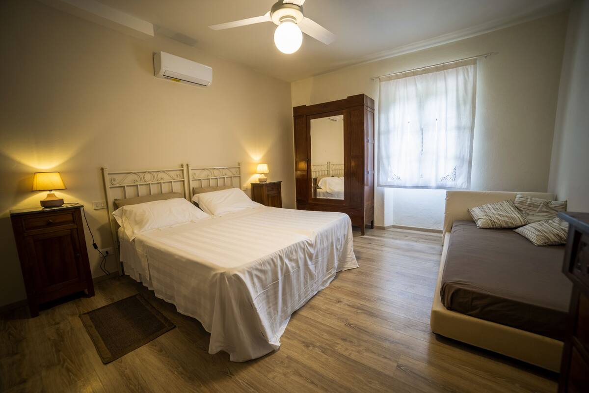 A bedroom with a large bed, a smaller sofa, a nightstand with a lamp, a wooden floor, and a ceiling fan.