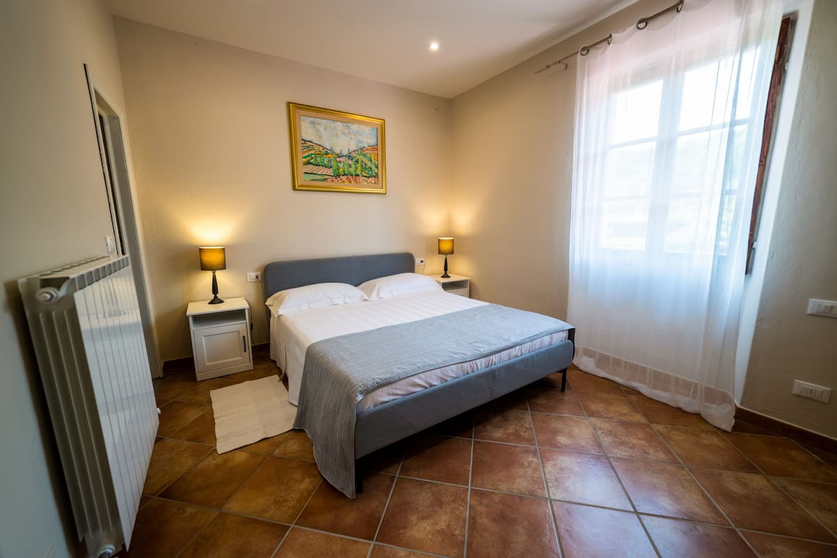 A bedroom with a bed, two bedside tables with lamps, a radiator, and a window with curtains.