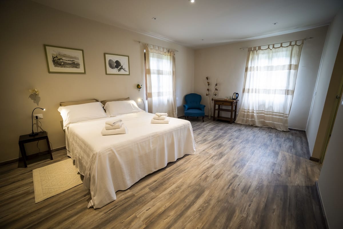 A bedroom with a large bed, a chair, a small table, and two windows with curtains.