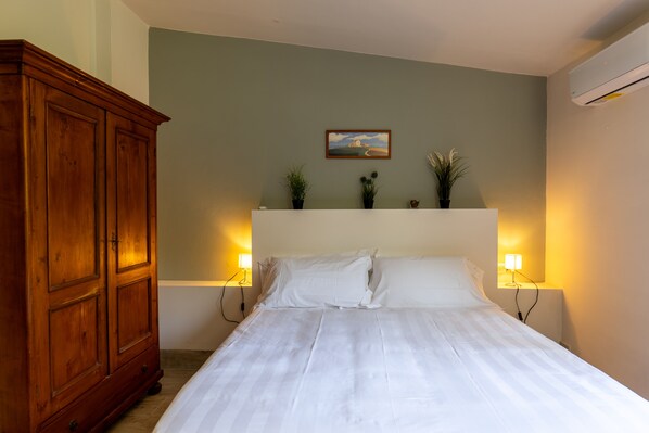 A bedroom with a large wooden wardrobe, a bed with white bedding, two bedside lamps, and a small wall-mounted shelf with plants.