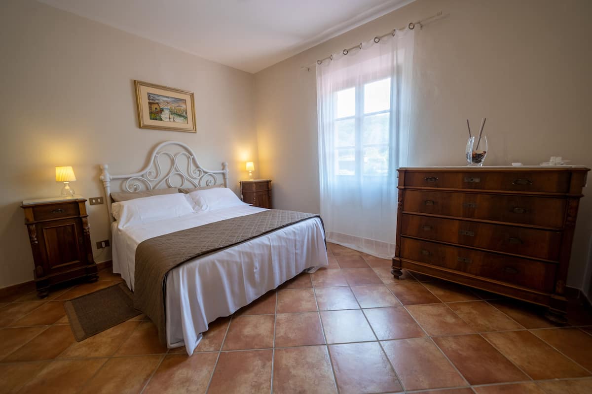 A bedroom with a bed, nightstands, a dresser, a painting on the wall, and a window with curtains.