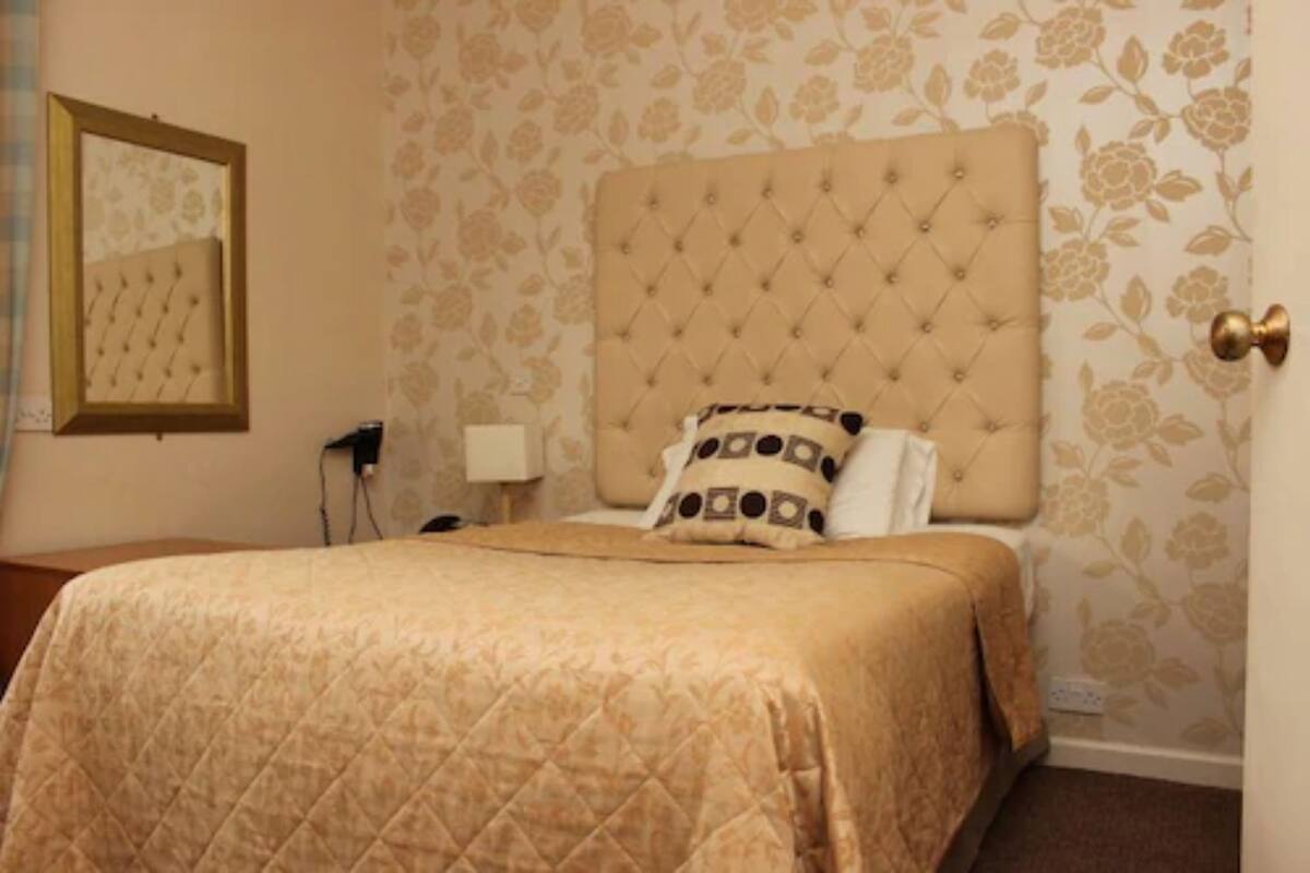 A hotel room with a large bed, a tufted headboard, floral wallpaper, a framed mirror, and a bedside table with a lamp.