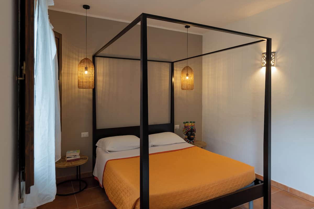 Ein Himmelbett mit weißer und orangefarbener Bettdecke, zwei Hängelampen, ein kleiner runder Tisch mit einem Buch und ein dekoratives Element auf dem Nachttisch.