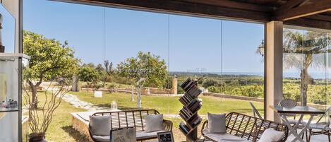 A covered outdoor seating area with wicker furniture, a glass table, and a view of a landscaped garden and distant landscape.