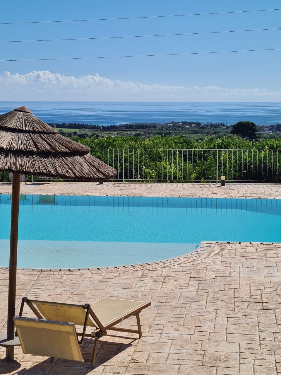 A swimming pool with a thatched umbrella and a lounge chair, surrounded by a stone patio and a fence.