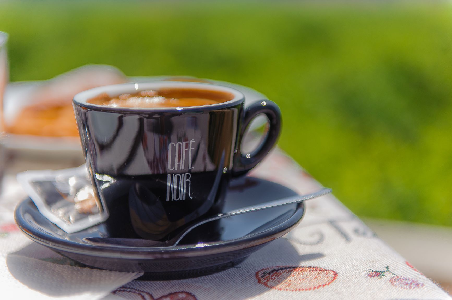 Una taza negra con el texto "CAFE NOIR" sobre un platito, colocada sobre una mesa con mantel estampado.