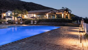 A swimming pool with lounge chairs and umbrellas, a covered outdoor seating area, and a hillside backdrop.