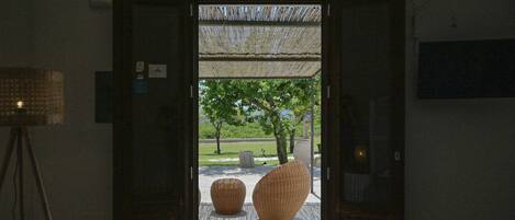 Een open deur die leidt naar een buitenruimte met een patroon vloer, twee grote geweven stoelen en een blauwe mat.