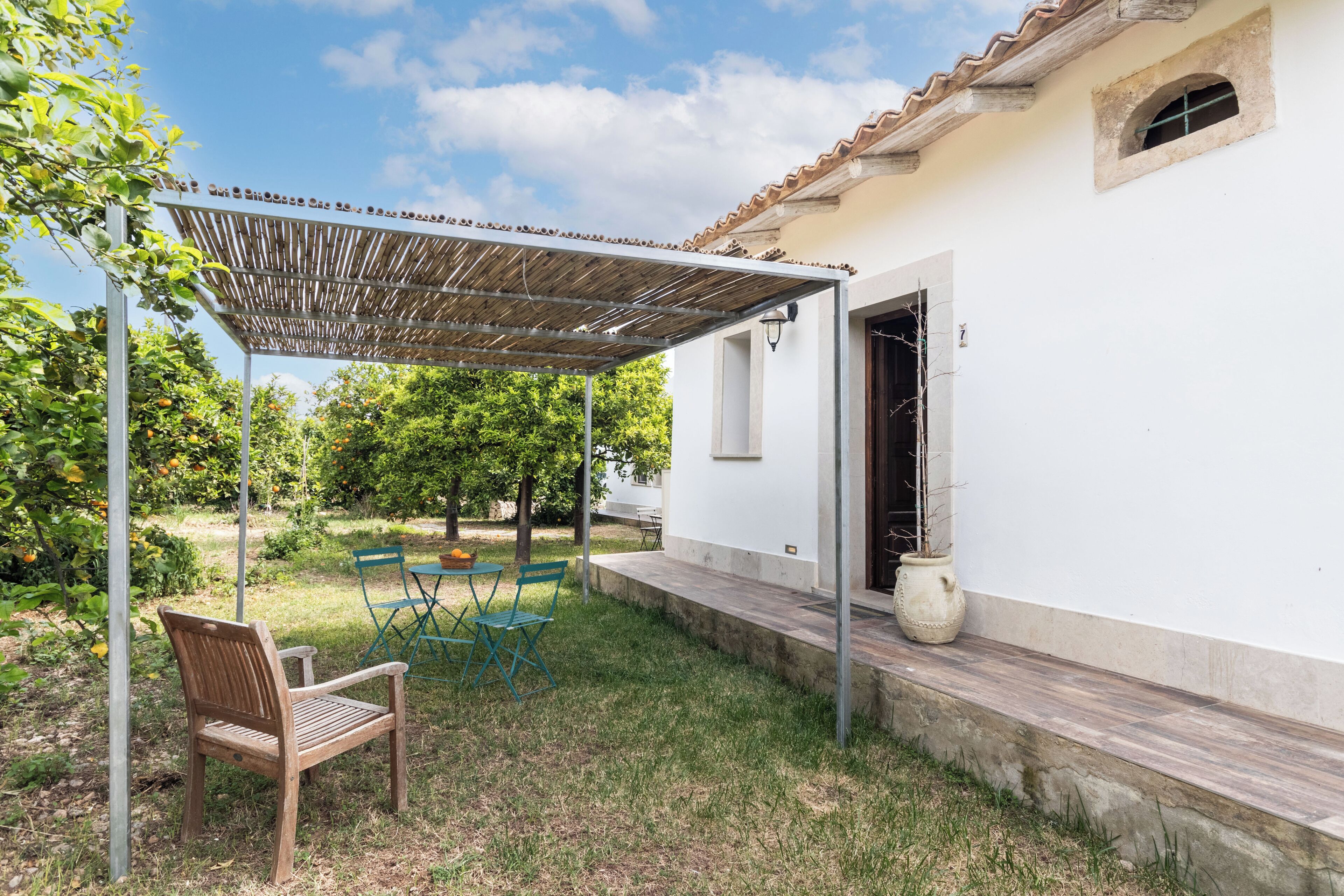 Un patio couvert avec un banc en bois et des chaises vertes, un bâtiment blanc avec un toit en tuiles, et une plante en pot sur le côté.