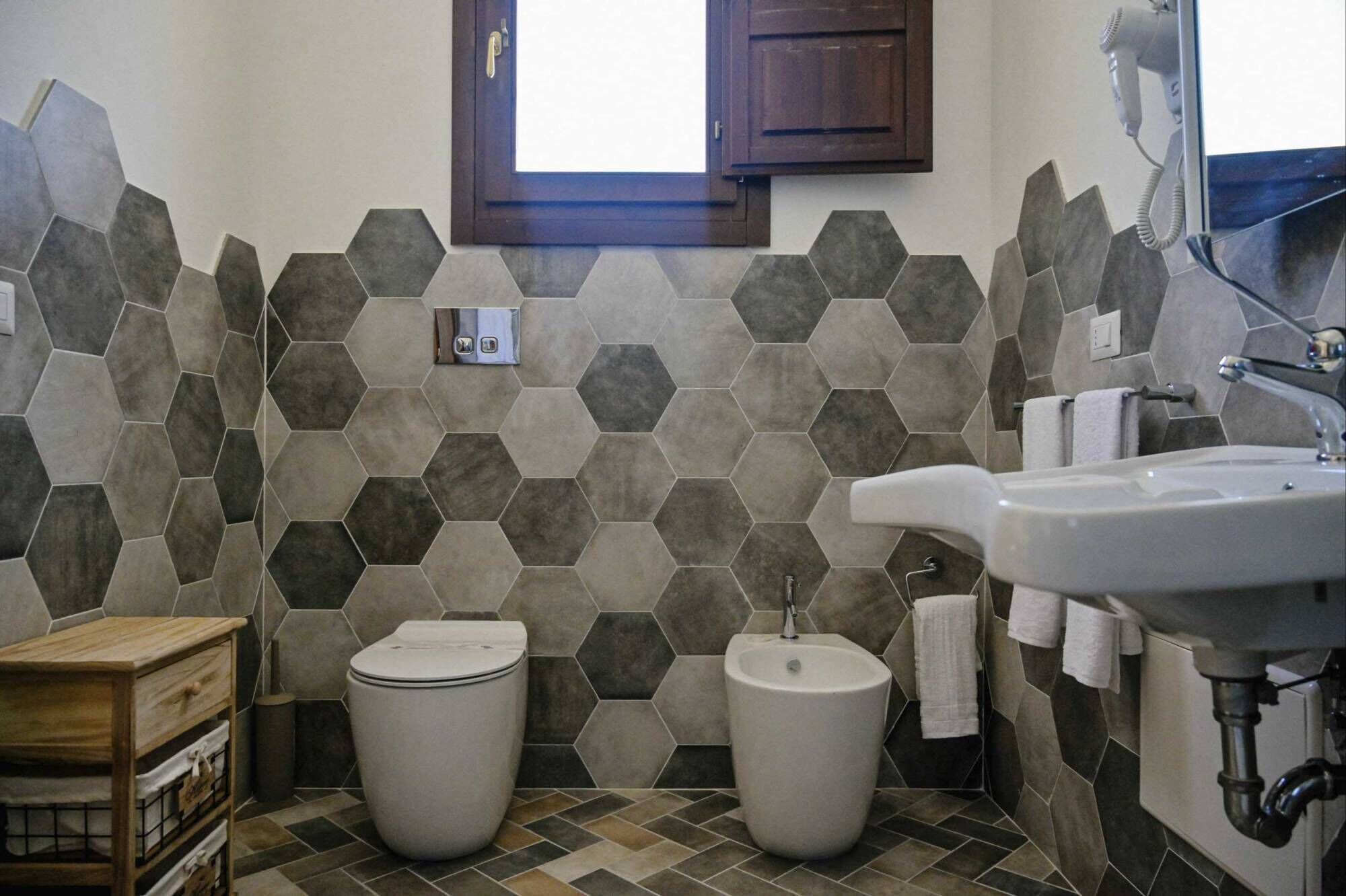 A bathroom with hexagonal patterned tiles, a toilet, a bidet, a sink, and a wooden cabinet.