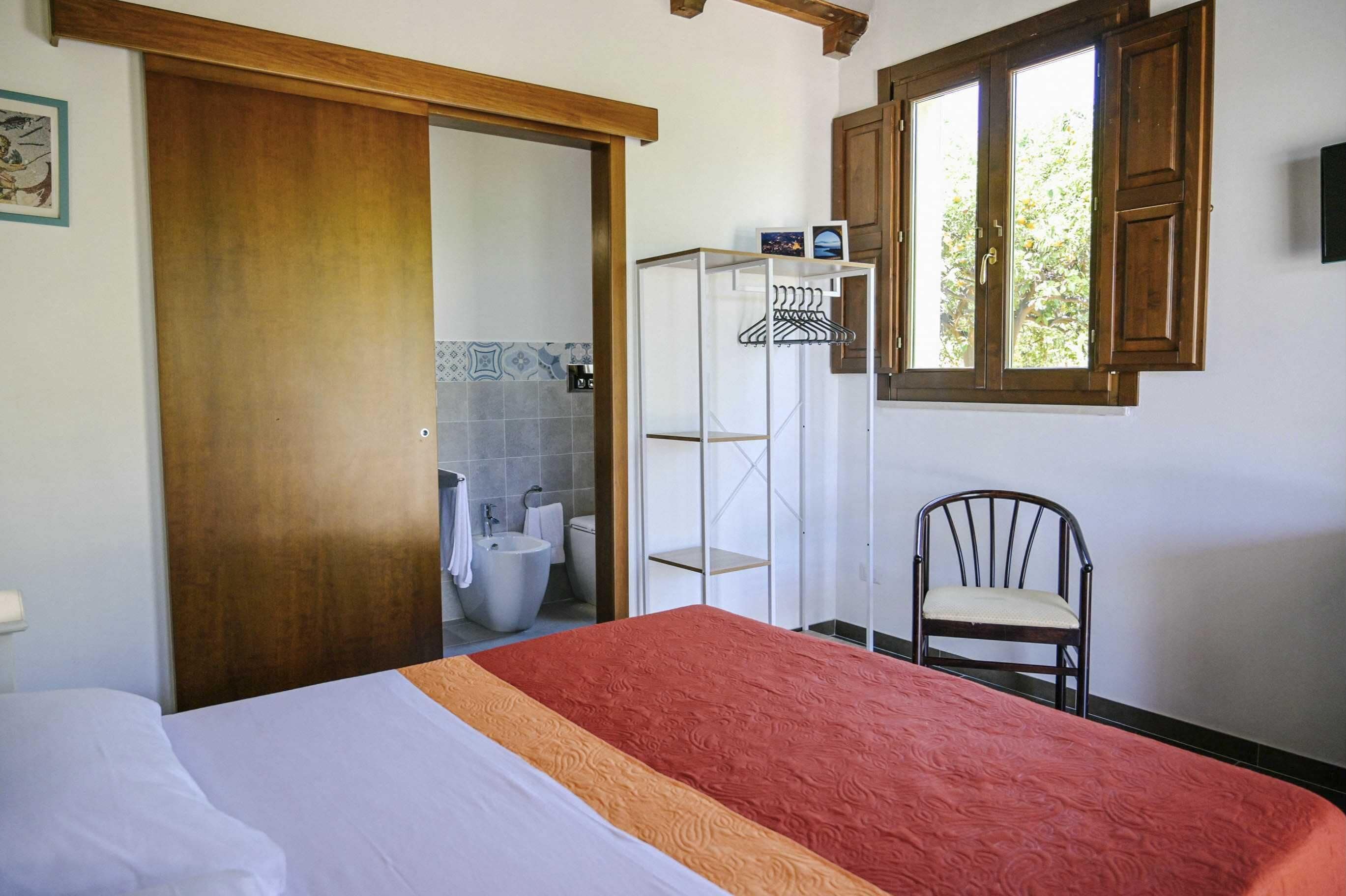 Hotellihuone, jossa on sänky, tuoli, puuovi, ikkuna ja kylpyhuone, joka näkyy avoimesta ovesta.