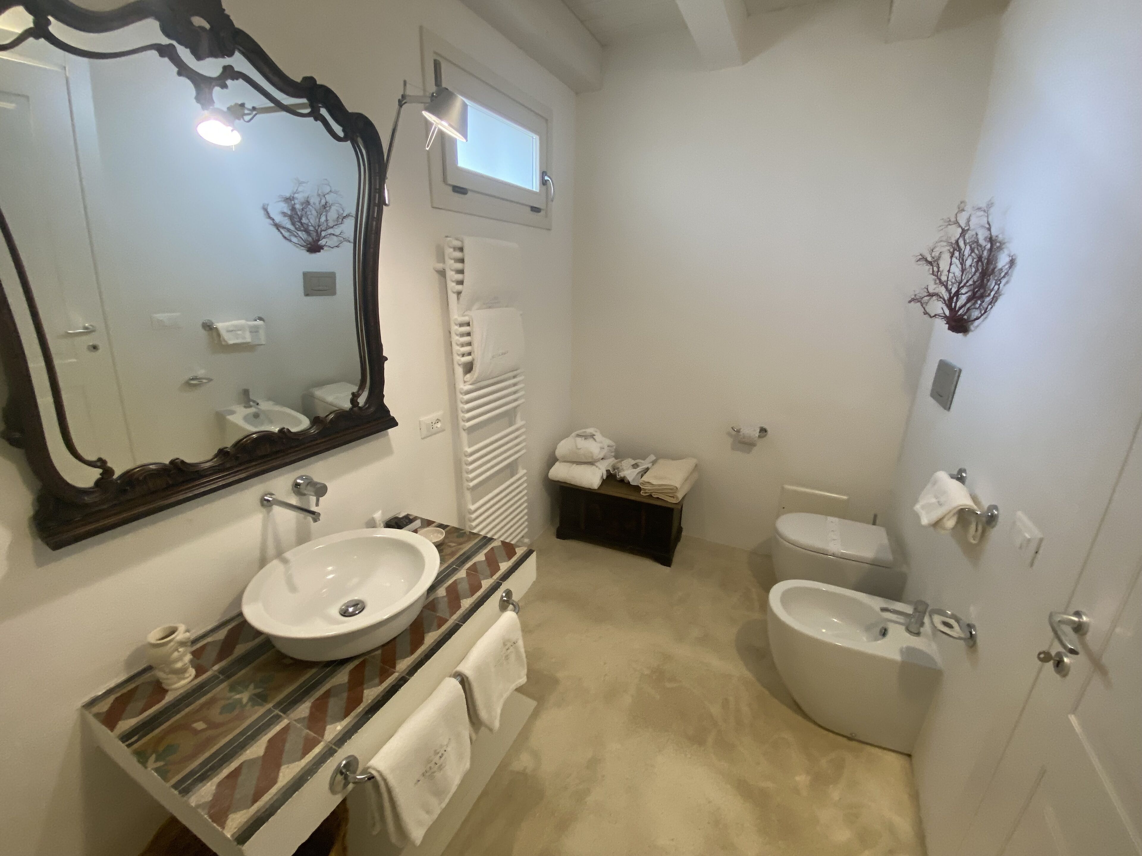 A bathroom with a large mirror, a sink, a toilet, and a bidet. There is a towel rack and a small shelf with toiletries.