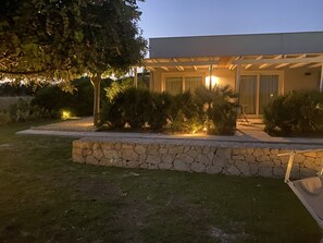 A modern house with a stone retaining wall, a covered patio, and a well-lit garden area. - Scilla Maris Charming Suites-Restaurant (Noto)