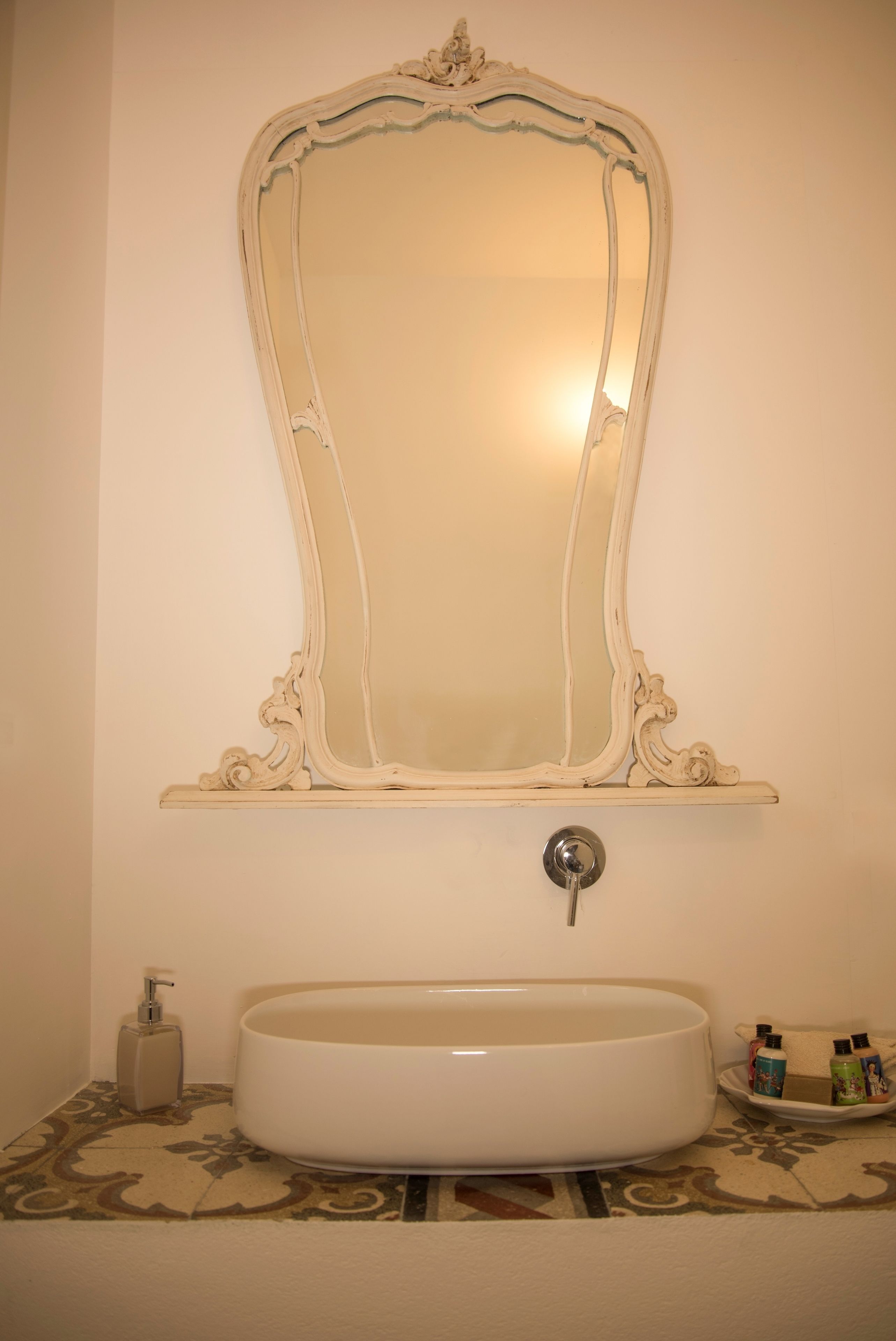 Une salle de bain avec un grand miroir décoratif, un lavabo blanc et une petite étagère contenant des articles de toilette.
