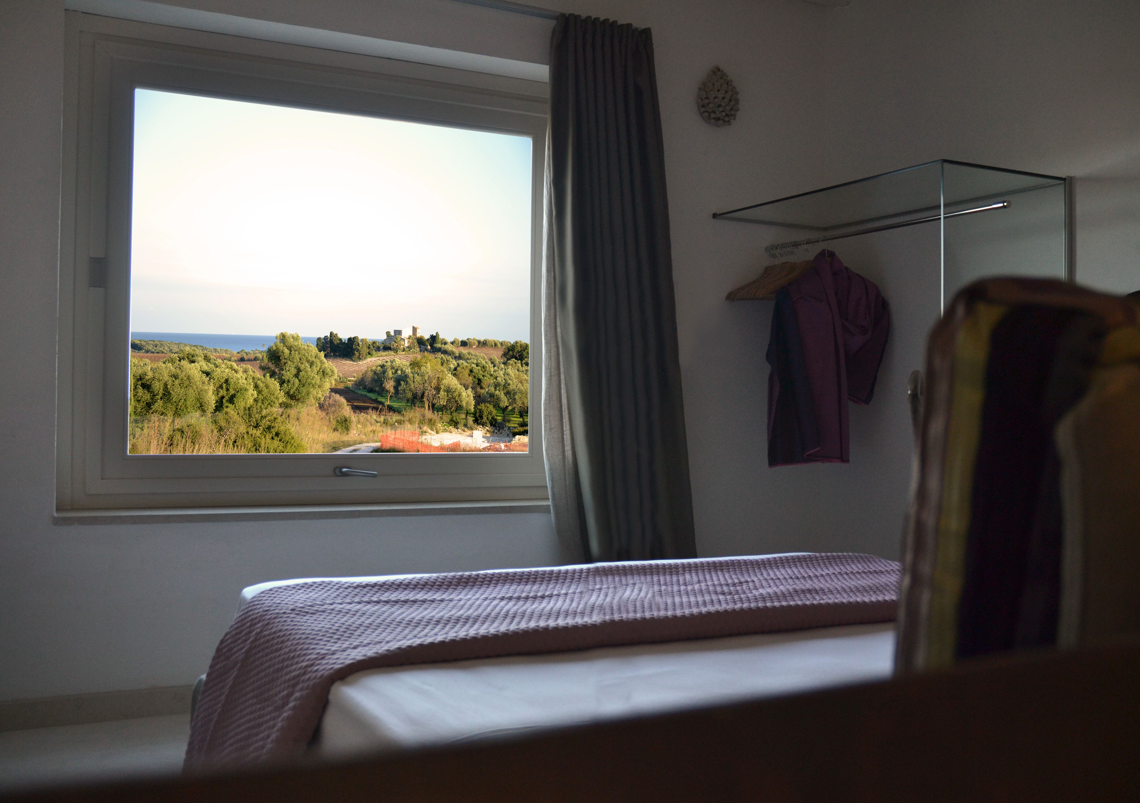 A bedroom with a large window, a bed, a hanging rack with clothes, and a view of a green landscape.