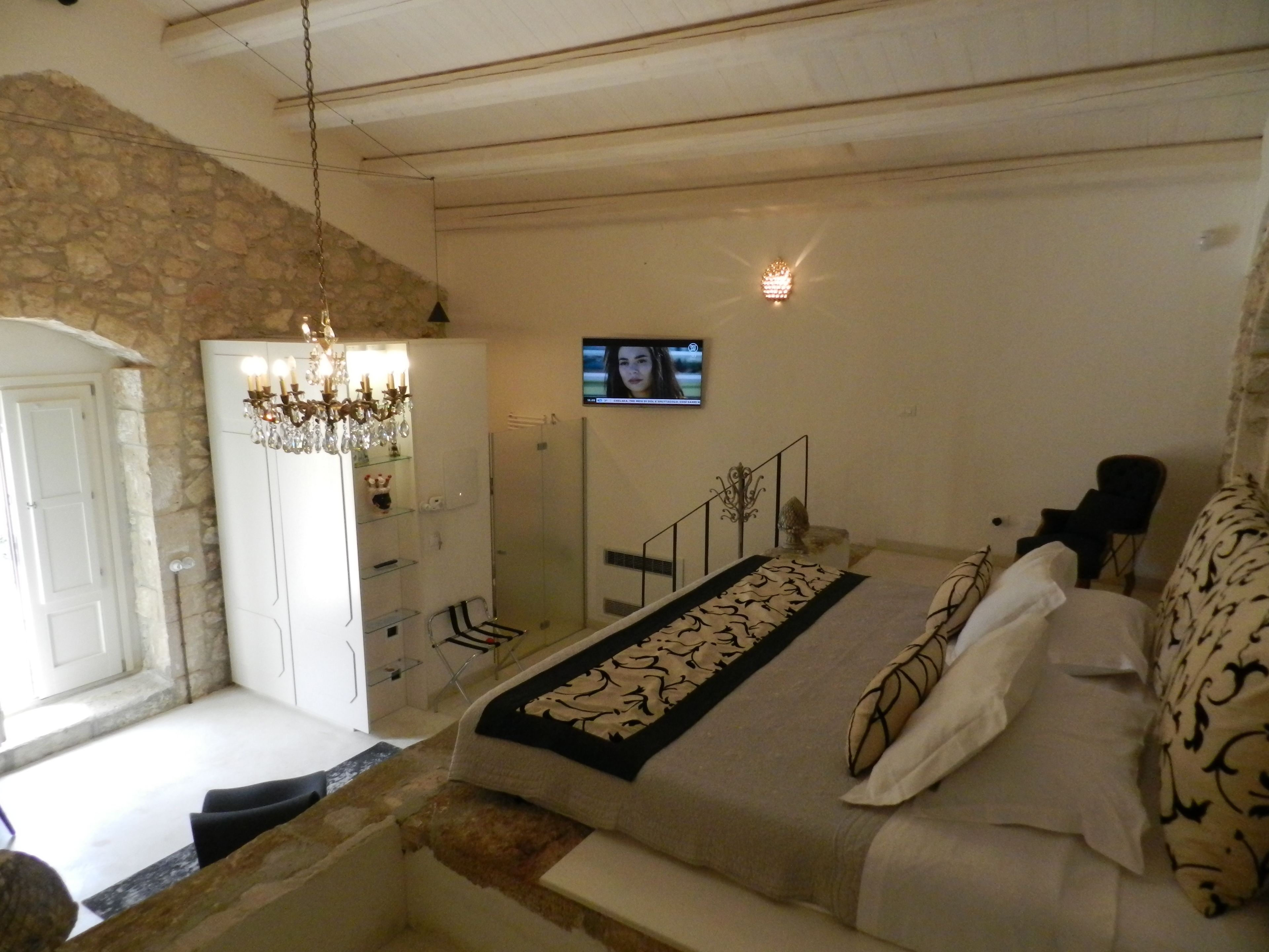 A bedroom with a stone wall, a chandelier, a TV, a bed with patterned bedding, and a staircase.