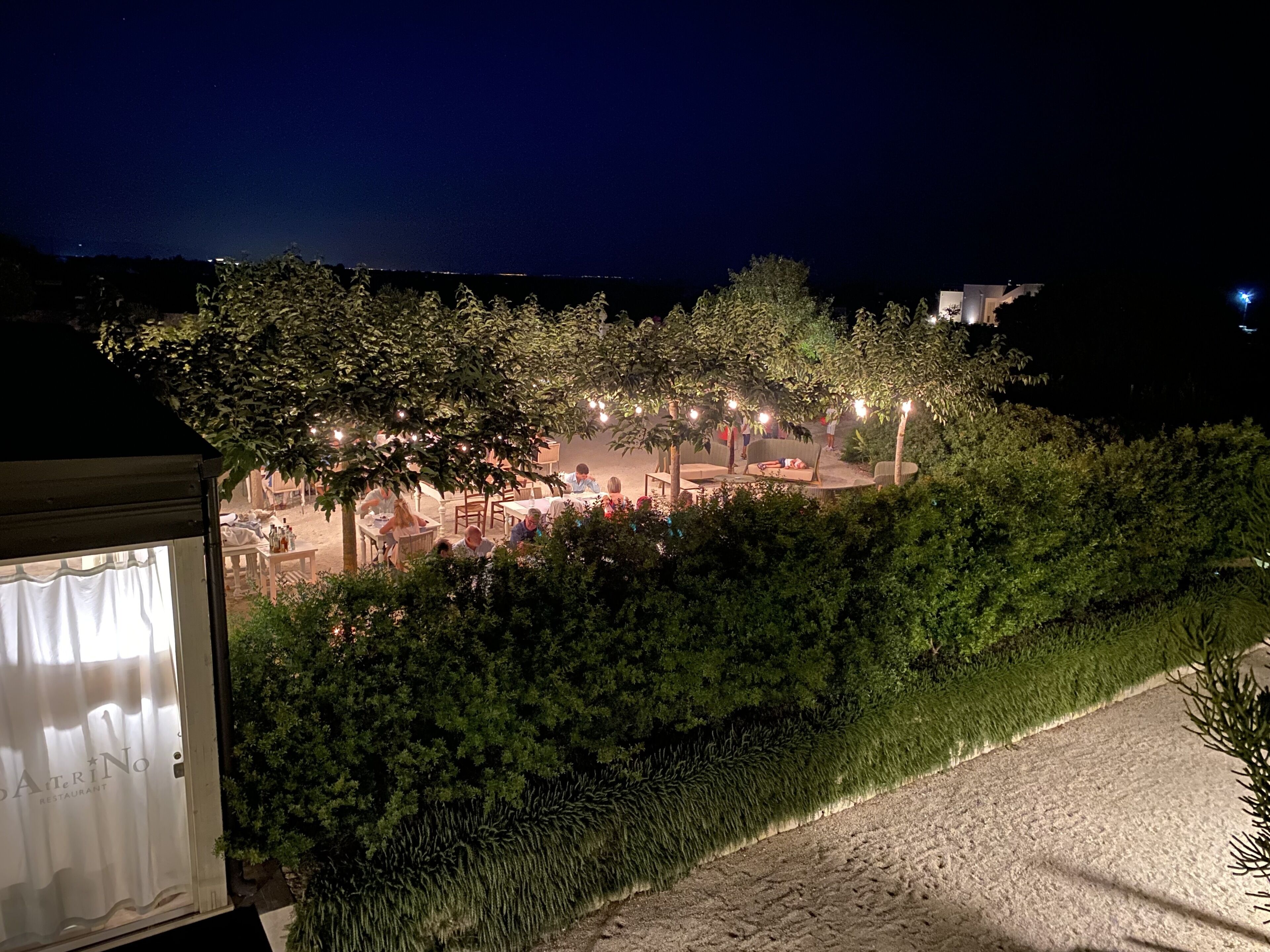 Un espace extérieur aménagé pour les repas en soirée, avec des tables et des chaises, entouré d’arbres et illuminé par des guirlandes lumineuses.