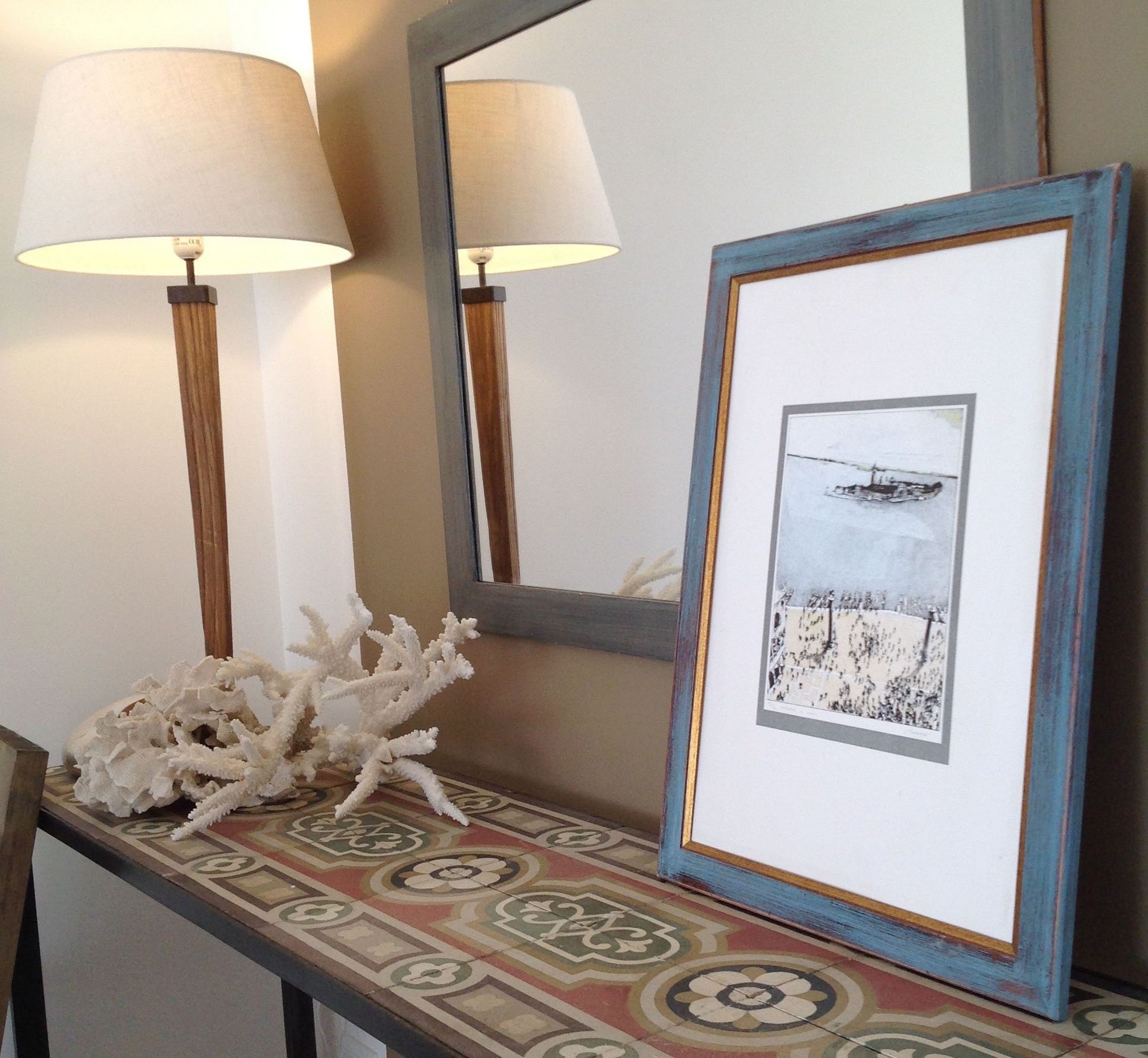 Une photographie encadrée représentant un paysage avec un bateau, une lampe et un objet décoratif en forme de corail, le tout posé sur une table à motifs.