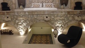 Un dormitorio con un arco de piedra, una cama con ropa de cama estampada, una araña y dos sillas negras.