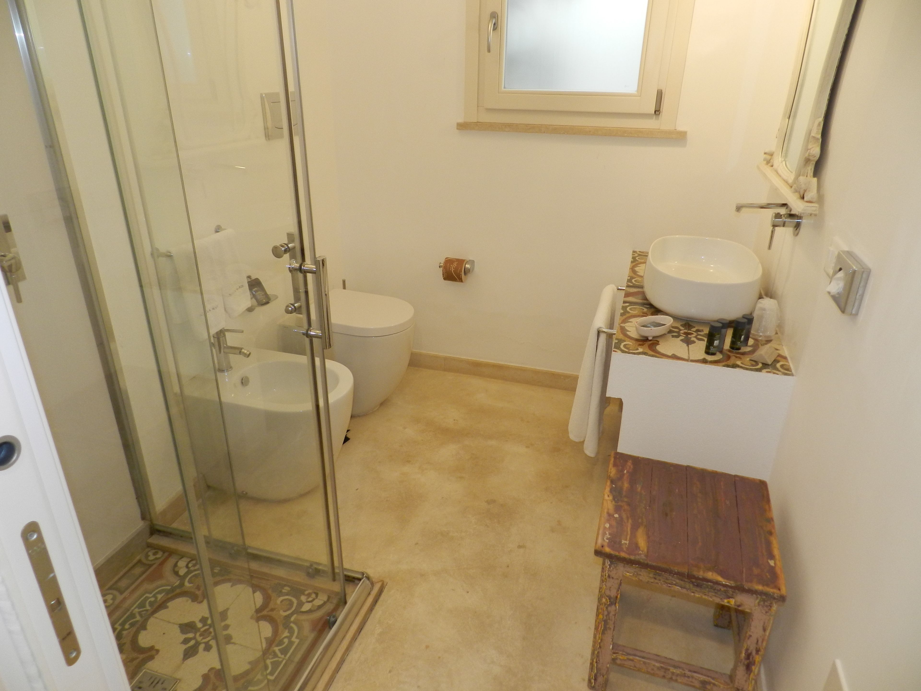 Un baño con cabina de ducha de vidrio, inodoro, lavamanos y un taburete pequeño de madera.