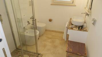 Un baño con cabina de ducha de vidrio, inodoro, lavamanos y un taburete pequeño de madera.