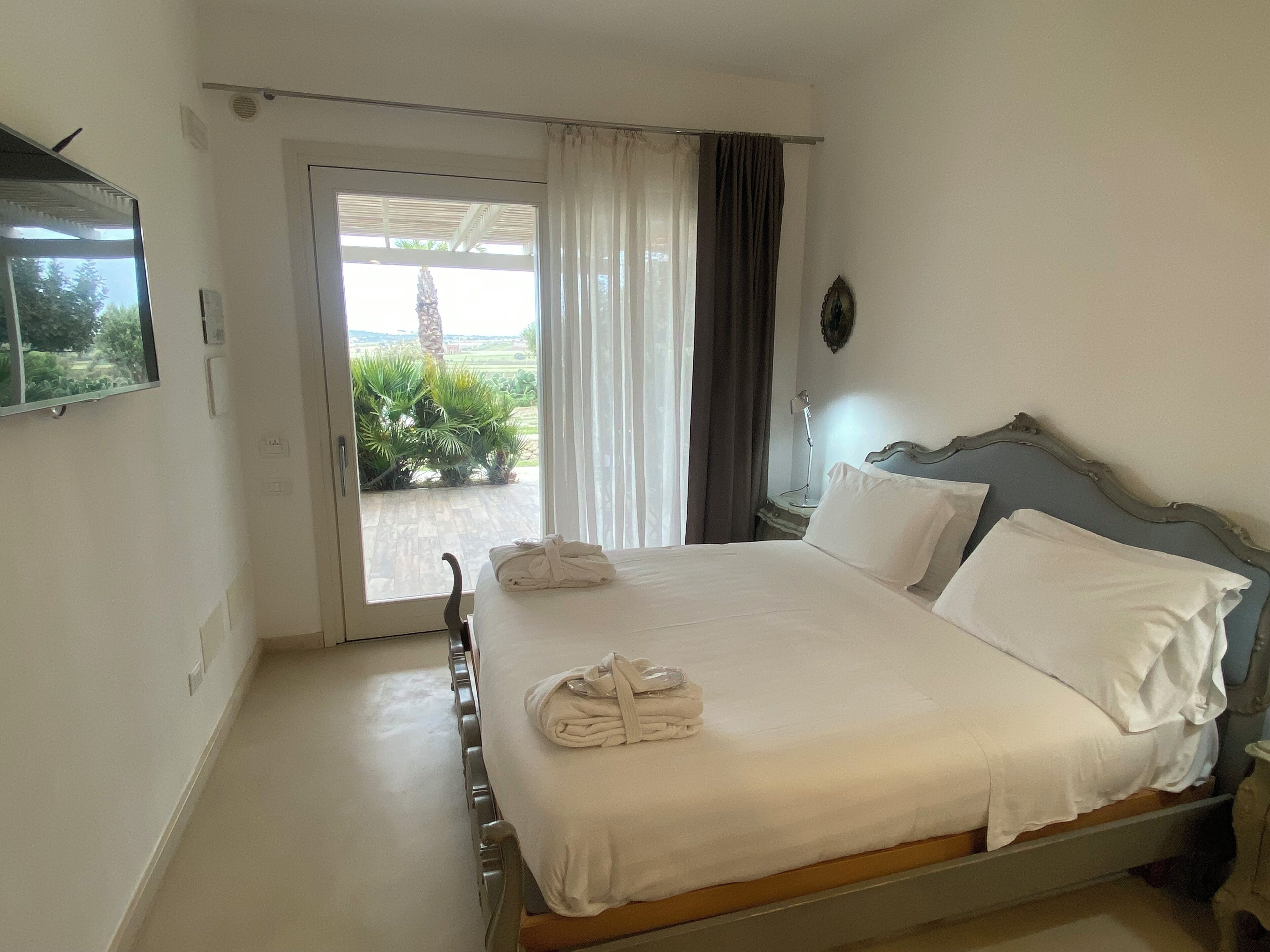 Habitación de hotel con una cama, televisor montado en la pared, ventana con cortinas y vista a zonas verdes.