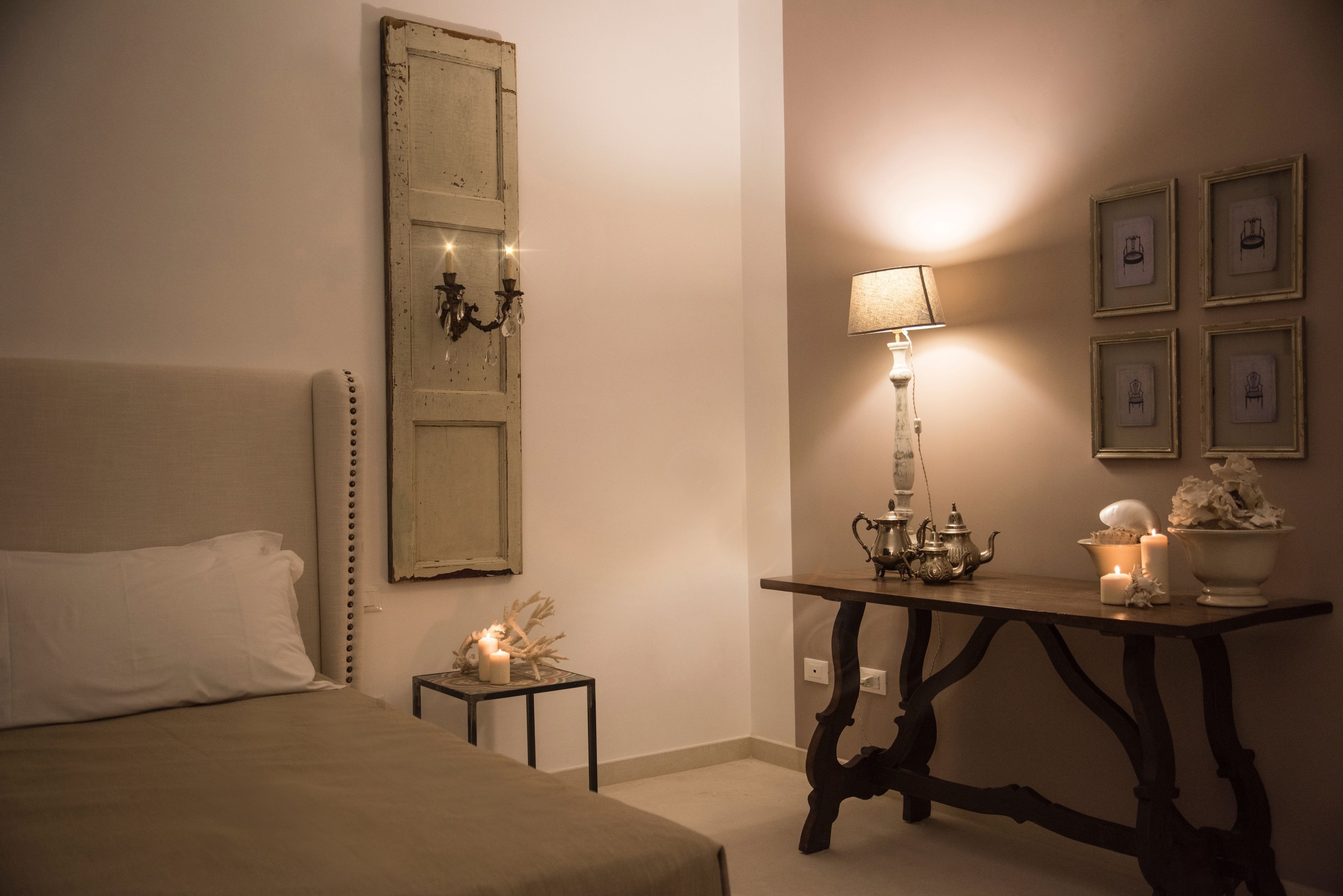 A bedroom with a bed, a nightstand with a lamp, a table with decorative items, and a wall with framed pictures.