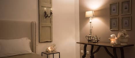 A bedroom with a bed, a nightstand with a lamp, a table with decorative items, and a wall with framed pictures.