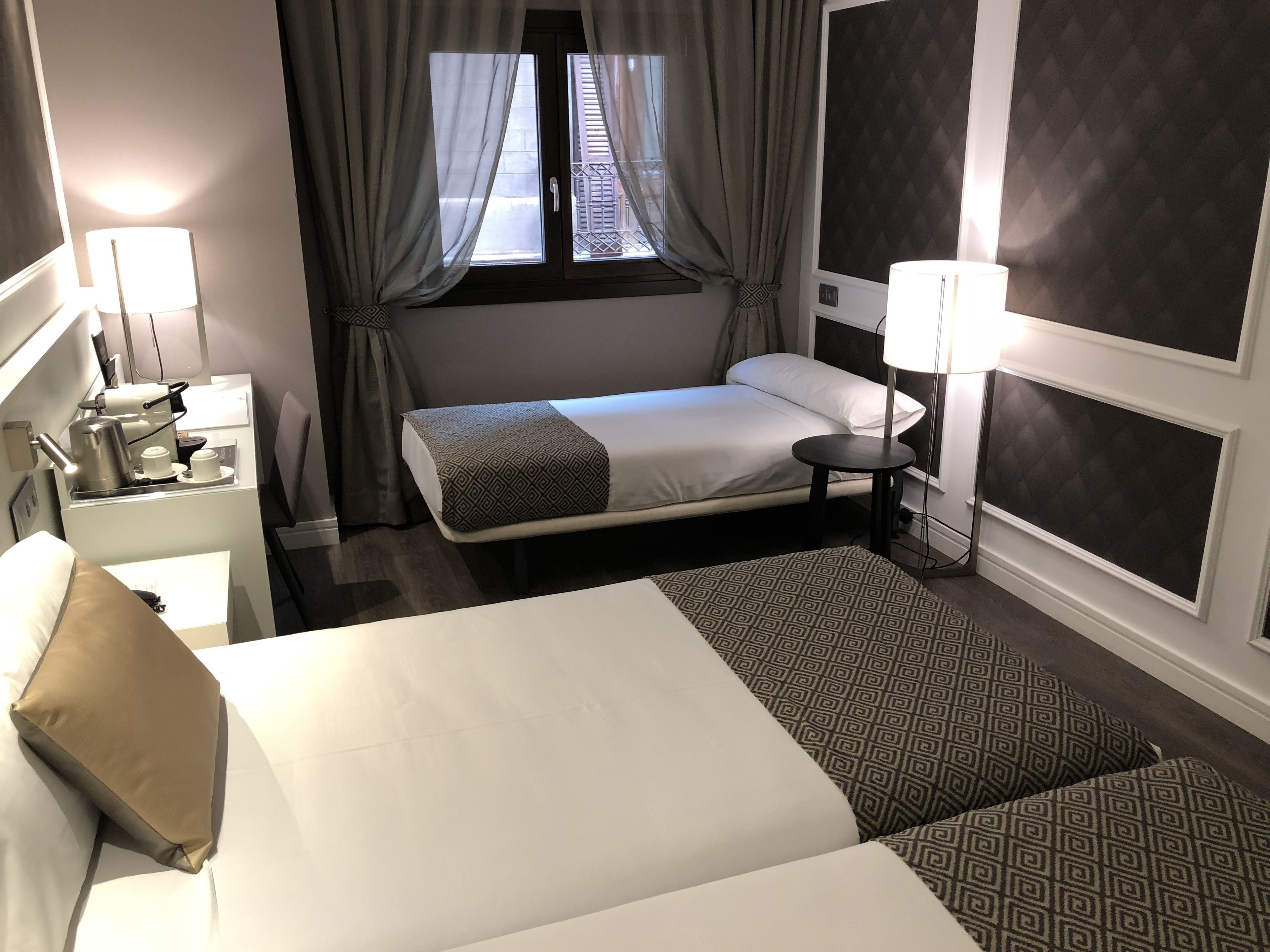 A hotel room with a large bed, a bedside table with a lamp, a small table, and a window with curtains.