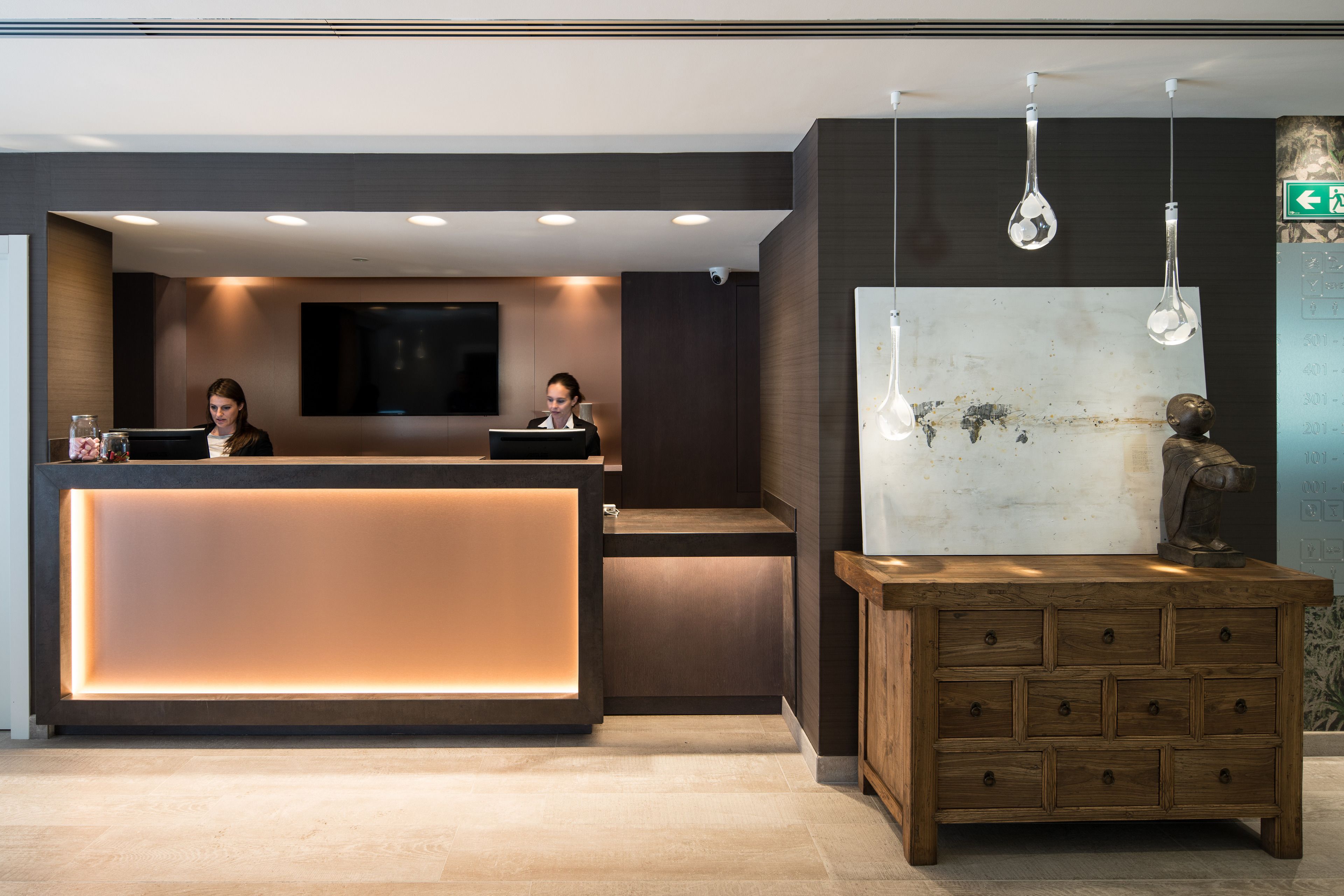 A reception desk with two staff members, a large wall-mounted TV, and a wooden cabinet with drawers.