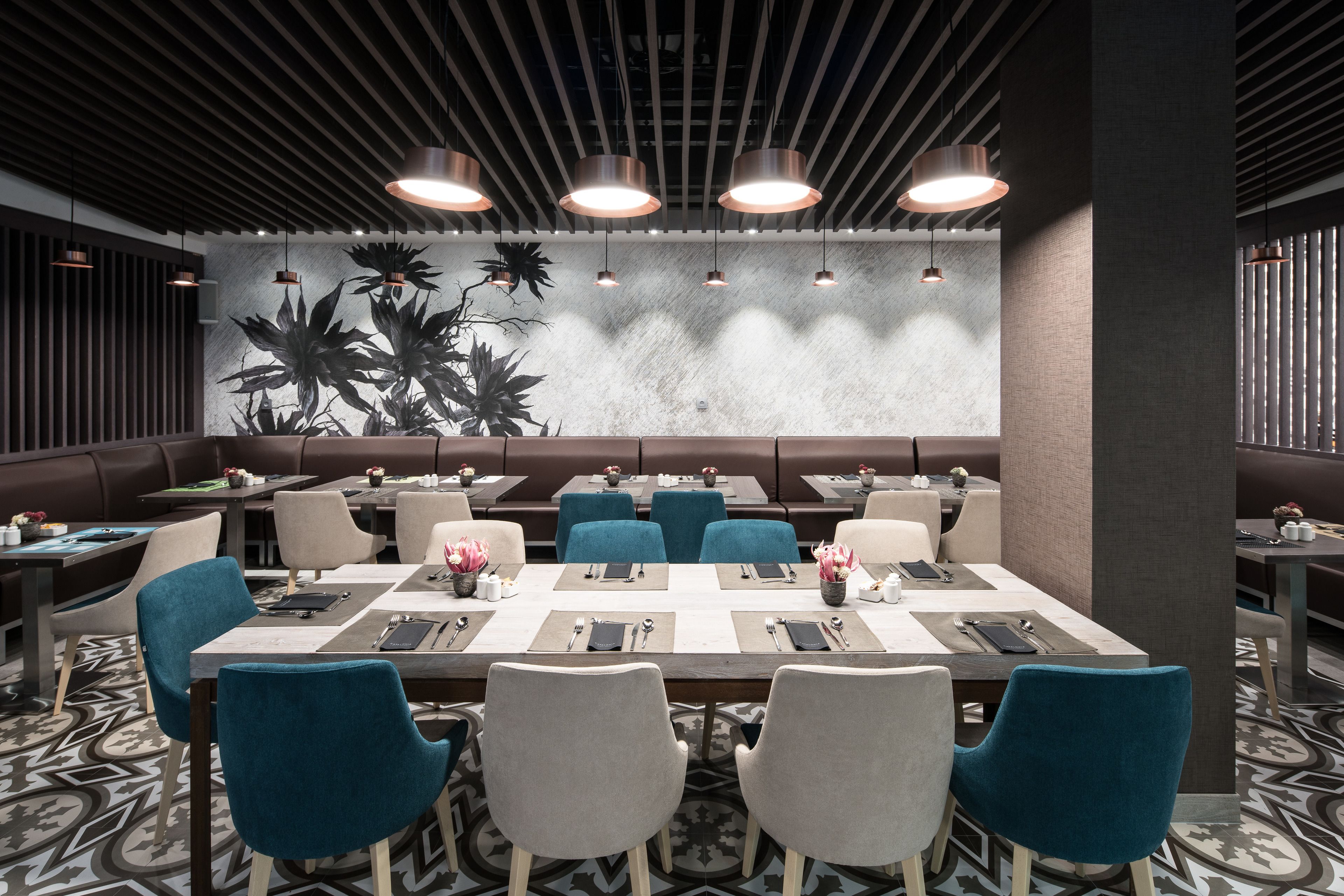 A modern restaurant with a long communal table, blue and grey chairs, and pendant lighting.