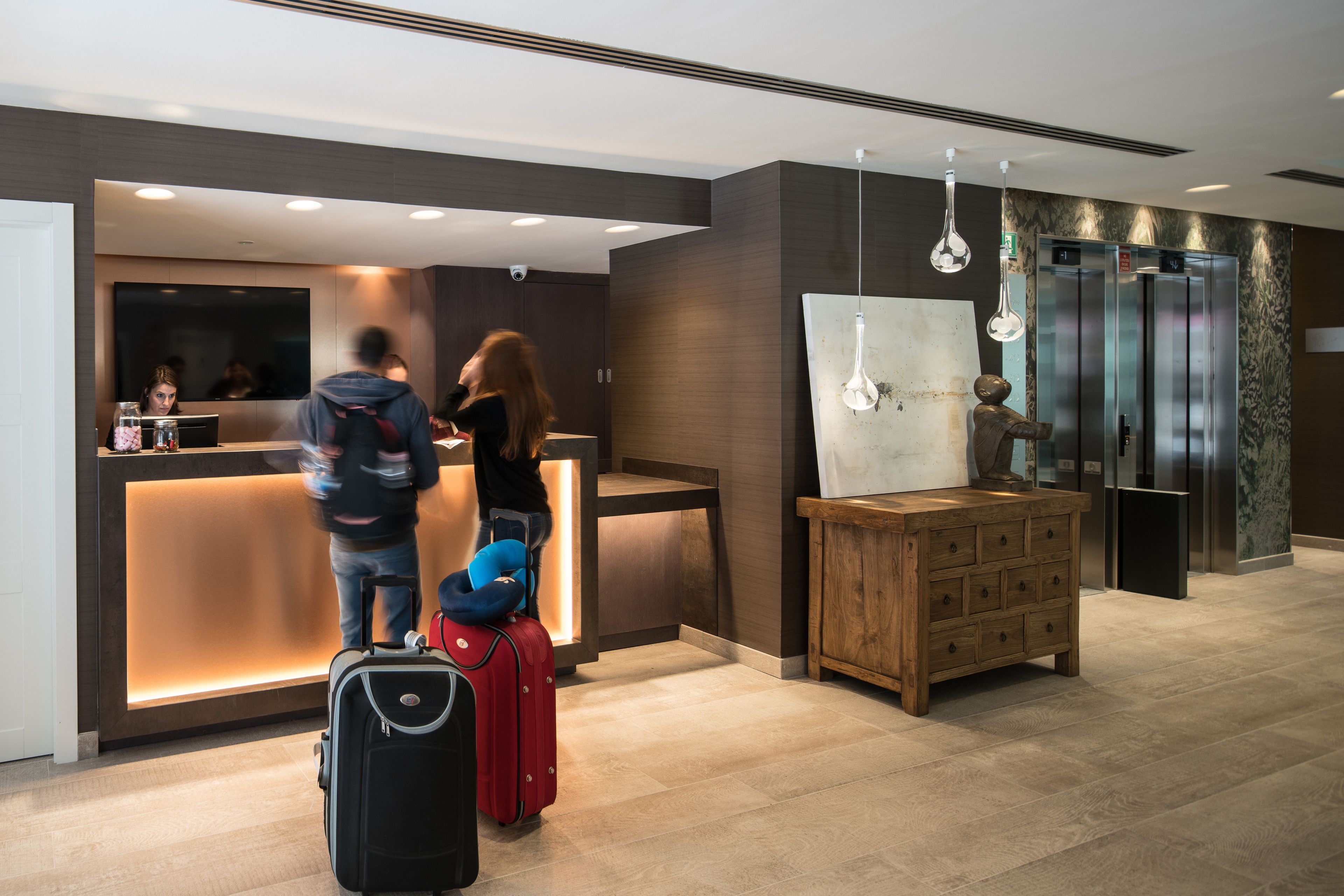 Ein Hotel-Rezeptionsbereich mit modernem Design, darunter ein Empfangstresen, ein Holzschrank und ein großer Spiegel. Im Vordergrund stehen zwei Koffer, und eine Person steht in der Nähe des Empfangstresens.