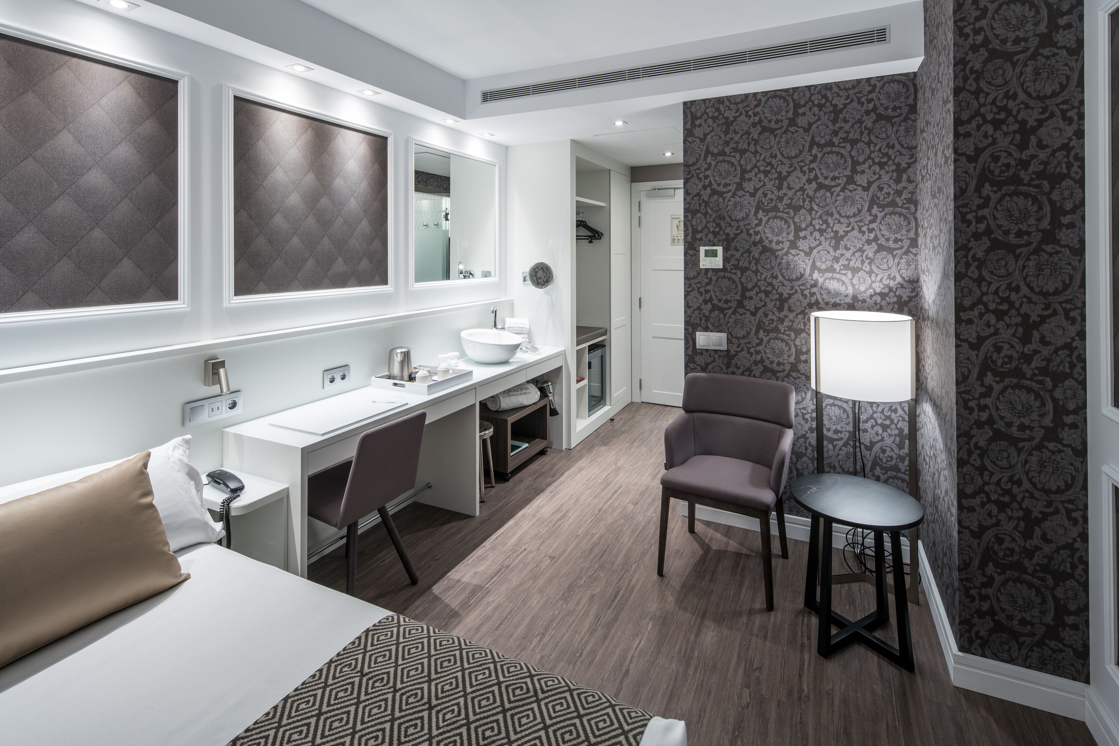 A hotel room with a bed, a desk with a sink, a chair, and a lamp.