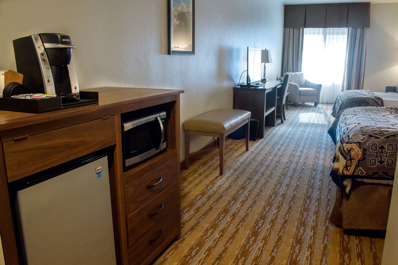 A hotel room with a kitchenette, a desk, a chair, a bed, and a window with curtains.