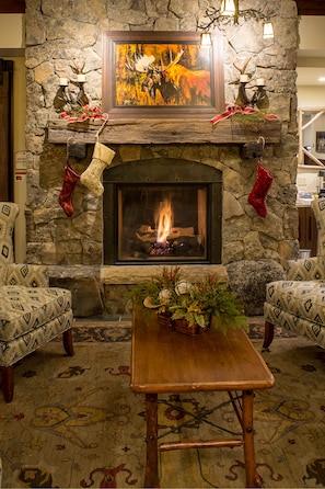 A stone fireplace with a fire, flanked by two patterned armchairs and a wooden table with a floral arrangement.