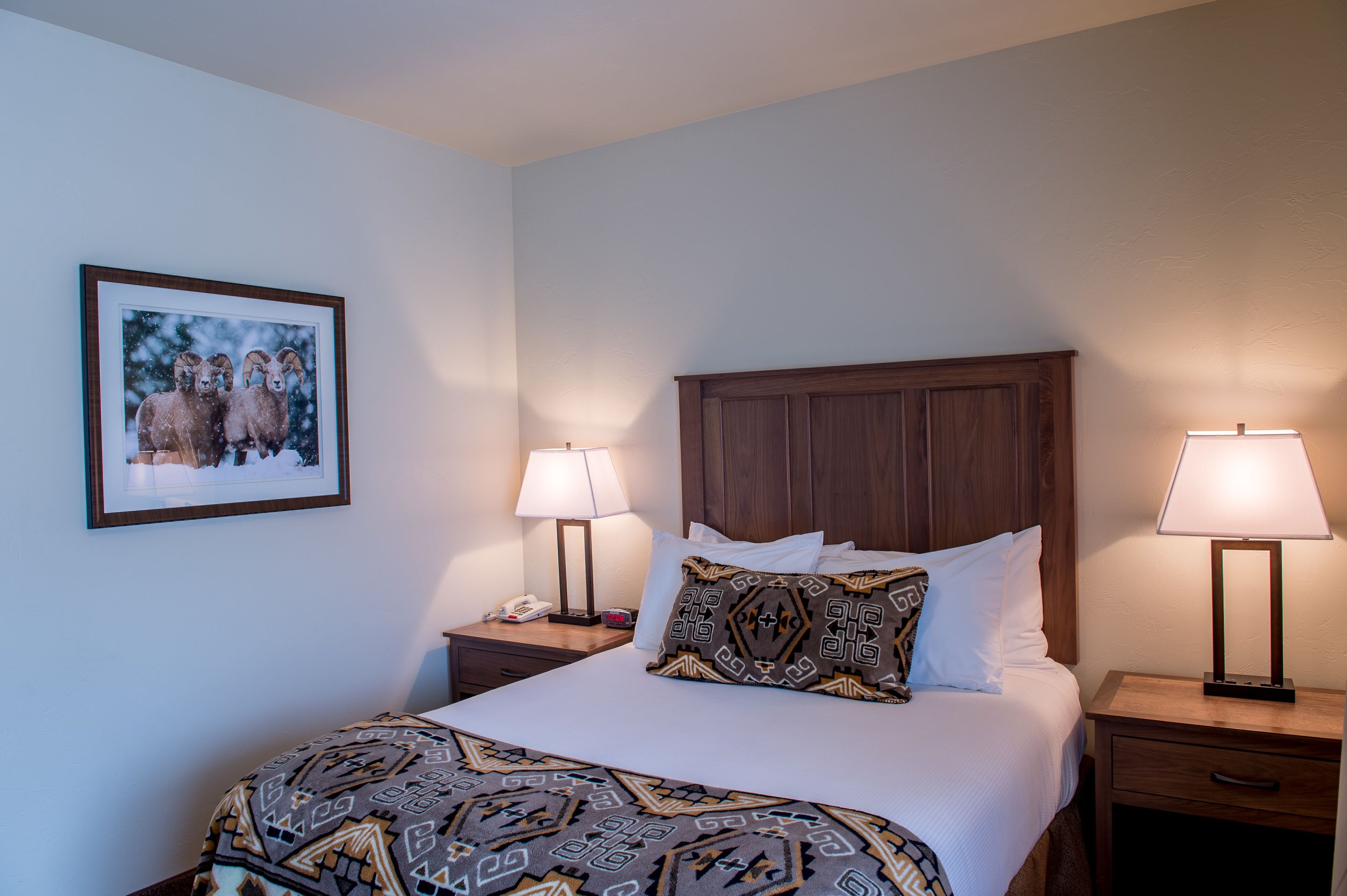 A neatly made bed with patterned bedding, two bedside lamps, a wooden headboard, a nightstand with a phone, and a framed picture on the wall.